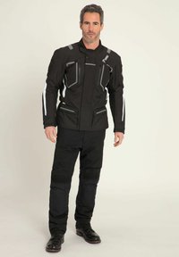 Czarna kurtka motocyklowa z białymi akcentami, kieszeniami zapinanymi na zamek i wysokim kołnierzem. Połączona z czarnymi spodniami z teksturowanymi panelami na kolanach i ciemnymi butami.