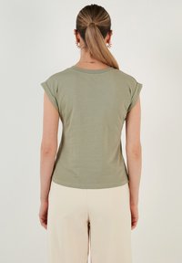Olivenfarvet kortærmet bluse med rund halsudskæring og oprullede ærmer. Glat tekstur, tætsiddende design og buet kant. Sættes sammen med lyse bukser.
