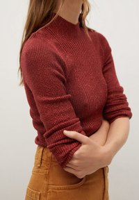 Henkilö, jolla on päällään ribbitetty viininpunainen pitkähihainen neule, jonka hihat on rukattu ylös, yhdistettynä korkeavyötäröisiin sinapinkeltaisiin housuihin.