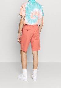 Maglietta a maniche corte con motivo tie-dye nei colori blu e arancione, abbinata a pantaloni corti corallo. I pantaloni corti presentano una texture liscia e tasche posteriori. Sneakers bianche.