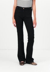 Femme portant un jean noir taille haute à jambes droites, un haut rayé noir et blanc, et des chaussures plates imprimé léopard, debout devant un fond blanc.