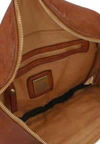 L'interno della borsa in pelle marrone presenta una cerniera dorata, una fodera in tessuto color nocciola e un'etichetta con testo. Include una piccola tasca con zip e un dettaglio elastico.