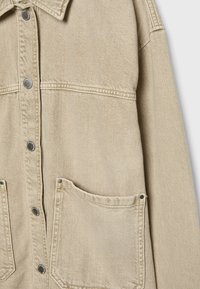 Veste en denim beige avec un devant à boutons, de grandes poches plaquées et un tissu texturé. Des accessoires en métal argenté soulignent le design.