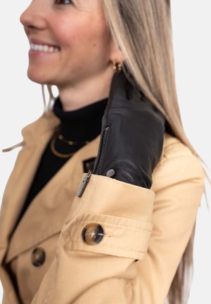 Schwarzer Lederhandschuh mit Reißverschlussakzent, getragen zu einem hellbeigen Trenchcoat mit runden Knöpfen und einem strukturierten Kragen.