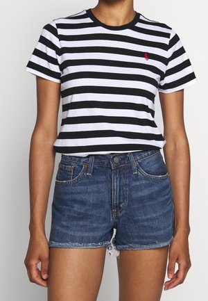 Person iført en sort og hvid T-shirt med vandrette striber og rød logo samt højtaljede blå cowboyshorts med frynsete kanter.