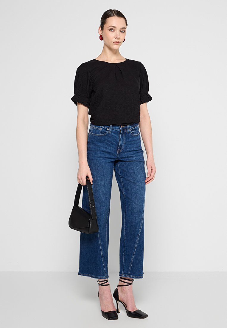 Schwarzes Kurzarmoberteil mit Falten-Detail, kombiniert mit hochgeschnittenen blauen Jeans. Das Model hält eine kleine schwarze Handtasche. Trägt schwarze Riemchen-Absätze.