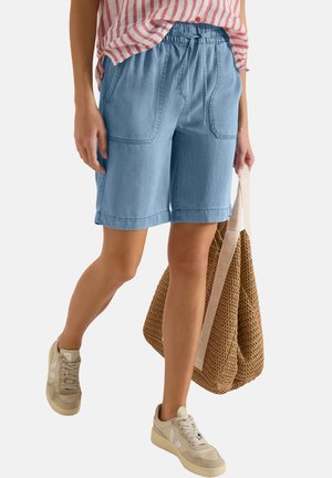 Frau trägt hellblaue knielange Shorts, gestreiftes rosa-weißes Oberteil, beige Turnschuhe und trägt eine braune, geflochtene Tragetasche.