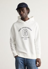 Sudadera blanca con un estampado gráfico frontal que dice "GANT SPORTSWEAR", combinada con una gorra azul oscuro. Modelo llevando unos vaqueros azules. Textura suave.