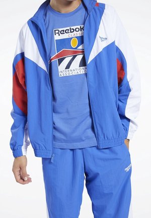 Person wearing blue, white, and red Reebok tracksuit with a blue Reebok Classic t-shirt showing a graphic logo on the front.