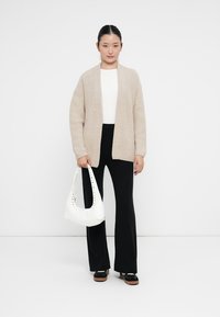 Gilet en tricot beige, top blanc, pantalon noir évasé et sac à main blanc avec détails cloutés. Chaussures décontractées avec une semelle marron.