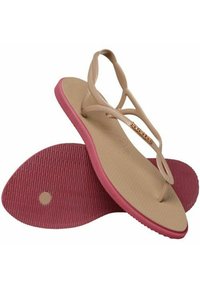 Tan flip-flop-sandaler med ribbede lyserøde såler. Har tynde, krydsede stropper med en metallisk detalje. Flad, fleksibel design for komfort.