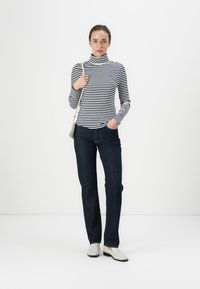 Pulover cu guler înalt, cu dungi negre și albe, purtat cu blugi de culoare albastru închis. Modelul poartă încălțăminte de culoare deschisă și ține o geantă mică argintie.