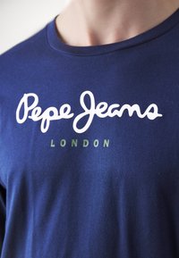 Sötétkék pamut póló, fehér 'Pepe Jeans' logóval és alul zöld 'LONDON' felirattal. Sima textúrájú és kerek nyakú design.