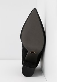 Semelle d'une chaussure noire à bout pointu avec un petit talon carré, positionnée contre un mur d'angle blanc et le sol.