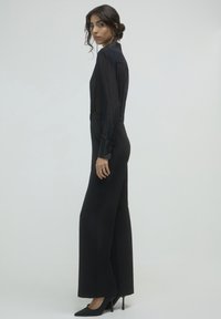 Femme portant un haut en voile noir à manches longues et un pantalon large taille haute noir, avec des talons noirs, se tenant de profil sur un fond clair.