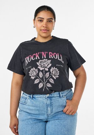 Mlada žena s tamnom kosom u crnoj "Rock 'N' Roll" majici s ružama, nosi plave prugaste traperice, stoji s rukom u džepu.