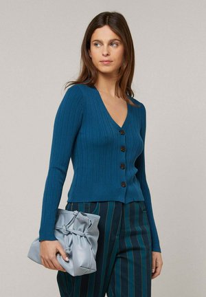 Frau trägt eine blaugrüne gerippte, geknöpfte Strickjacke und dunkelblaue, fein gestreifte Hose, hält eine hellgraue gerüschte Clutch-Tasche.