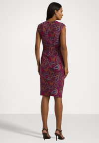 Vestido ajustado em roxo com padrão paisley. Mangas curtas e corte até ao joelho. Feito de tecido suave. Sandálias de salto alto mostradas.