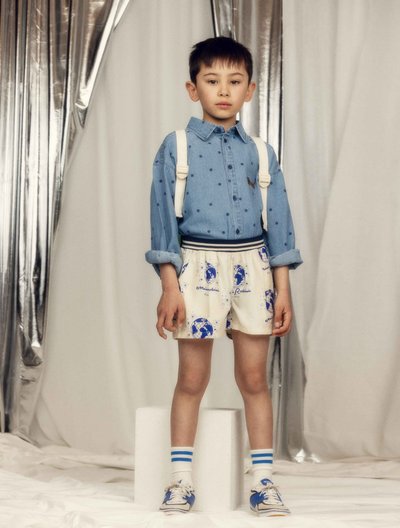 Νεαρός αγόρι που φοράει μπλουζάκι με μπλε πουά, κρεμ σορτς με μπλε σχέδια, άσπρο σακίδιο, ριγέ κάλτσες και μπλε αθλητικά παπούτσια, στέκεται μέσα σε κλειστό χώρο.