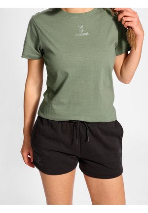 Femme portant un t-shirt hummel vert olive et un short noir à cordon, tenant ses cheveux tressés d'une main.