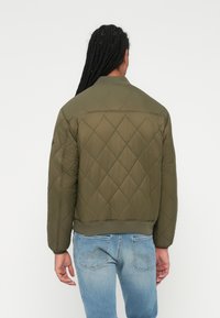 Oliven grøn quiltet jakke med ribbet krave, struktureret diamantmønster og elastiske manchetter, parret med lyseblå jeans.