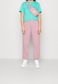 Persona con camiseta de manga corta color verde azulado, pantalones deportivos rosas, zapatillas blancas y bolso cruzado rosa de Adidas, sobre un fondo liso.