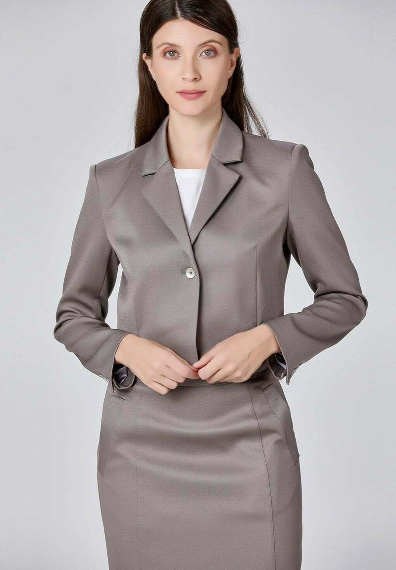 Blazer ajusté gris avec une fermeture à un bouton, des revers accentués et des épaules structurées. Texture lisse, associé à un haut blanc.