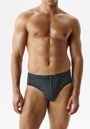 Umięśniony mężczyzna w czarnych, wzorzystych bokserkach z etykietą "Atlantic", stojący na jednolitym białym tle.