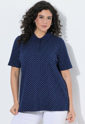 Kobieta z kręconymi włosami, ubrana w granatową koszulkę polo z krótkim rękawem ozdobioną małymi, wielokolorowymi kropeczkami oraz białe spodnie na białym tle.