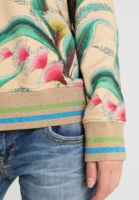 Sweatshirt med blommönster som har en beige bas, färgglada inslag i rosa och grönt, samt en texturerad, ribbad nederkant med horisontella ränder.