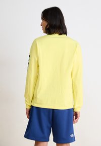 Chemise jaune à manches longues avec un col rond et une coupe décontractée, associée à un short bleu ; présente un motif coloré sur la manche gauche.