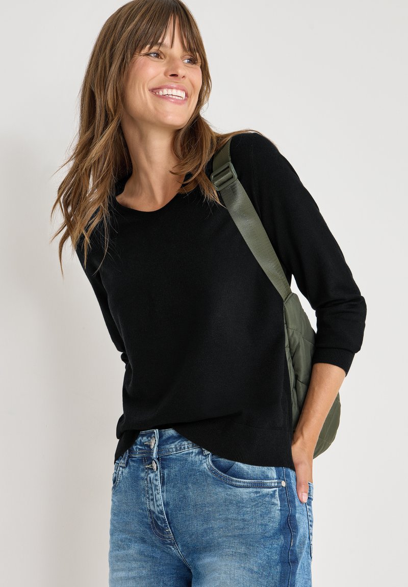 Maglione nero a maniche lunghe con scollo rotondo, abbinato a jeans in denim blu chiaro e una borsa a spalla verde imbottita.