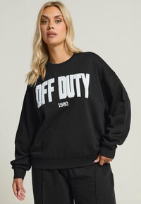 Czarna bluza z luźnym krojem, z wyraźnym białym napisem "OFF DUTY" oraz detalem "1980". Wykonana z teksturowanej tkaniny z ściągaczami na końcach rękawów.