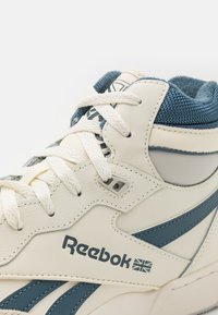 Högtoppsneakers i vitt läder med blå detaljer, med textilstruktur vid kragen, snörning och Reebok-logotypen på sidan.