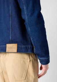 Tamna plava traper jakna s kožnom etiketom na kojoj piše "Calvin Klein Jeans." Bež hlače s velikim stražnjim džepovima i suptilnom teksturom.