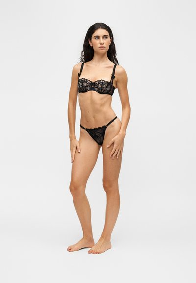 Femme debout pieds nus portant une lingerie noire en dentelle avec des motifs floraux sur un fond uni clair, regardant sur le côté.