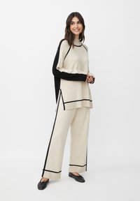 Beige Strickpullover und weit geschnittene Hosen mit schwarzen Akzenten. Der Pullover hat einen hohen Kragen und Seitenschlitze; weiche Textur über die gesamte Fläche.