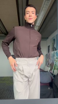 Young person wearing a brown zip-up jacket and high-waisted beige pants, standing indoors with artwork and bags in the background.