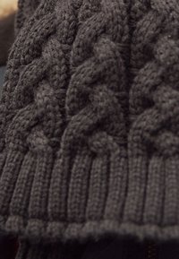 Tissu tricoté gris foncé avec un motif à gros câble épais et bord côtelé, probablement une partie d'un pull ou d'une écharpe.