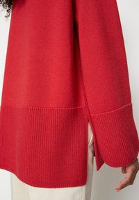 Pull rouge en maille avec une finition texturée, présentant des fentes sur les côtés et des manches longues. Détail côtelé notable à l'ourlet et aux poignets.