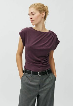 Mujer con cabello rubio recogido en un moño, vistiendo una camiseta sin mangas morada metida dentro de pantalones grises con cinturón negro, manos en los bolsillos, mirando hacia la izquierda.