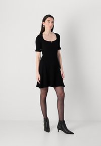 A woman stands confidently in a fitted black dress with short sleeves and a flared skirt, paired with black ankle boots.