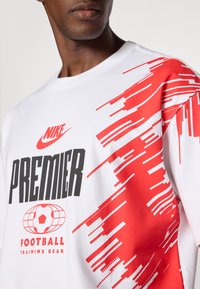 Hvid Nike T-shirt med stort sort "PREMIER" tekst, røde grafiske mønstre og et fodboldgrafik. Blødt bomuldsmateriale.