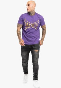 Lila pamut póló "TAPOUT" logóval sárgában; rövid ujjú, kerek nyakú, testhezálló fazon. Fekete, szakadt farmerral és fehér sportcipővel viselve.