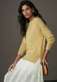 Pull en maille jaune avec des poignets et un ourlet côtelés, associé à une jupe en dentelle blanche à rayures verticales. Mannequin tenant un accessoire noir.