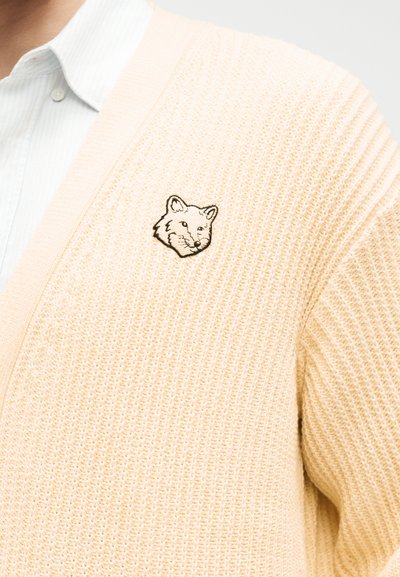 Κρεμ πλεκτή ζακέτα με μικρό κεντημένο κεφάλι αλεπούς στο αριστερό στήθος, φορεμένη πάνω από λευκό πουκάμισο με γιακά.