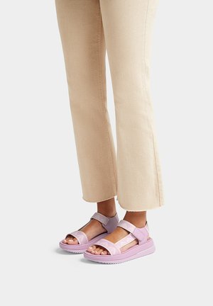 Paarse sandalen met een gestructureerde bovenkant, verstelbare banden en een gevoerde zool, gecombineerd met lichtbeige flared broek.
