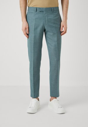 Szövetes, textúrált türkiz nadrág, slim fit szabással, éles redőkkel, oldalzsebekkel, fehér sportcipővel párosítva.