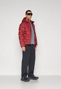 Röd vadderad jacka med texturerad yta, kombinerad med en grå t-shirt och mörka, löst sittande byxor. Svarta sneakers fulländar outfiten.