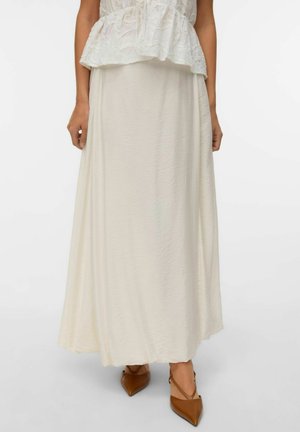 Žena v dlhej, prúdiacej, smotanovo bielej sukni a hnedých lodičkách so špicatou špičkou na jednoduchom svetlom pozadí.